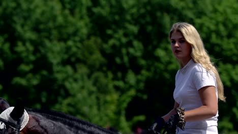 a beautiful girl in white hair and white clothes slowly rides on a black brown stallion. the girl finished training and goes to the recreation area. the girl is a little tired and sweat is visible on her face. sunny summer day on a green glade.