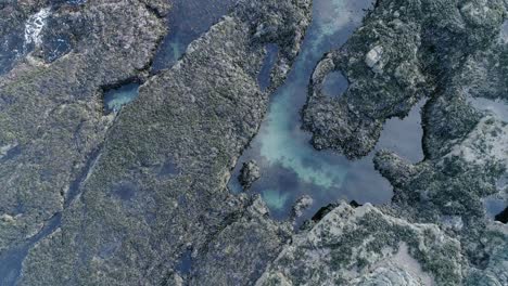 Rocky-craggy-coastal-rock-aerial-tracking-downwards-towards-a-rockpool-surrounded-by-seaweed-and-mossy-rock