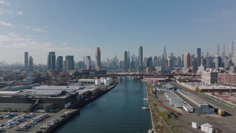 Forwards-fly-above-Newtown-Creek,-warehouses-and-logistic-sites-on-waterfronts.-Modern-business-skyscrapers-in-background.-New-York-City,-USA