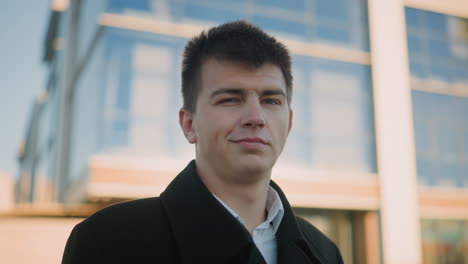 man in black clothing smiling confidently at camera with soft bright light illuminating from behind, background featuring reflective glass building