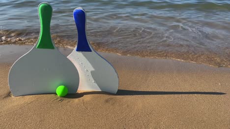 pair of beach tennis paddle rackets and green balls on sandy beach with waves breaking on shore