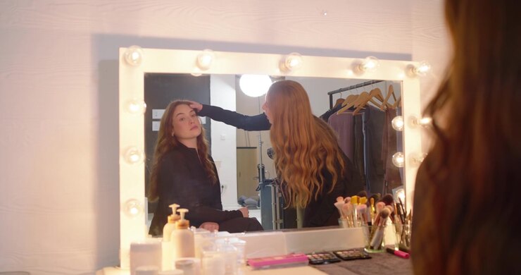 Female stylist leaning into vanity mirror reflection checking makeup with brushes, client watching