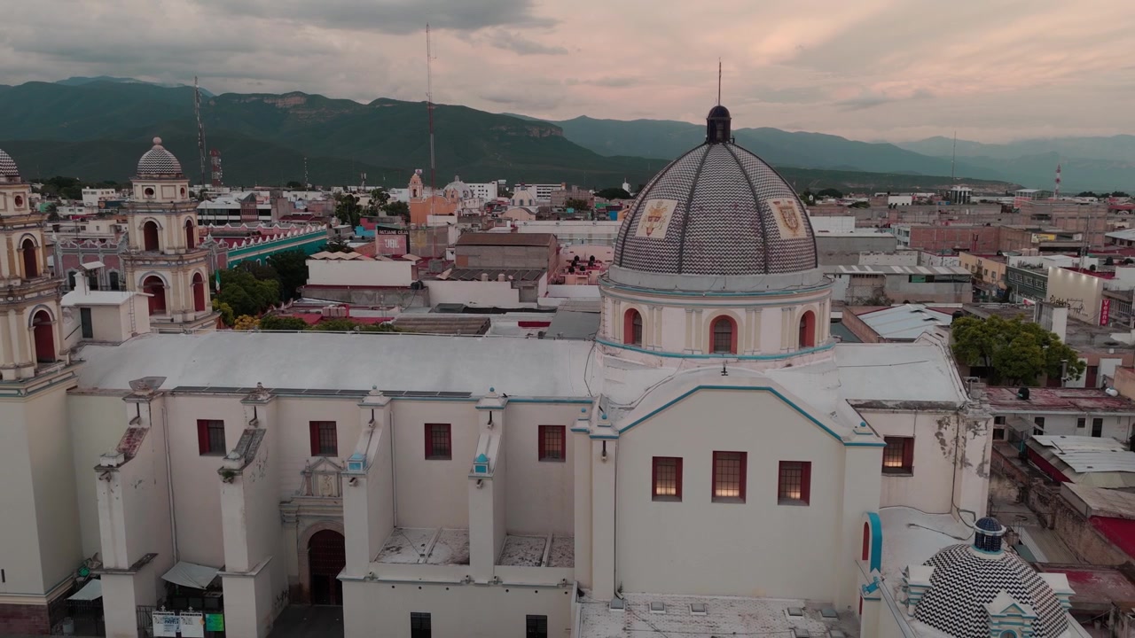 Vista aerea con drone che mostra una chiesa storica con tetto a cupola e campanili, circondata da edifici cittadini e catene montuose sotto un cielo nuvoloso in Messico.