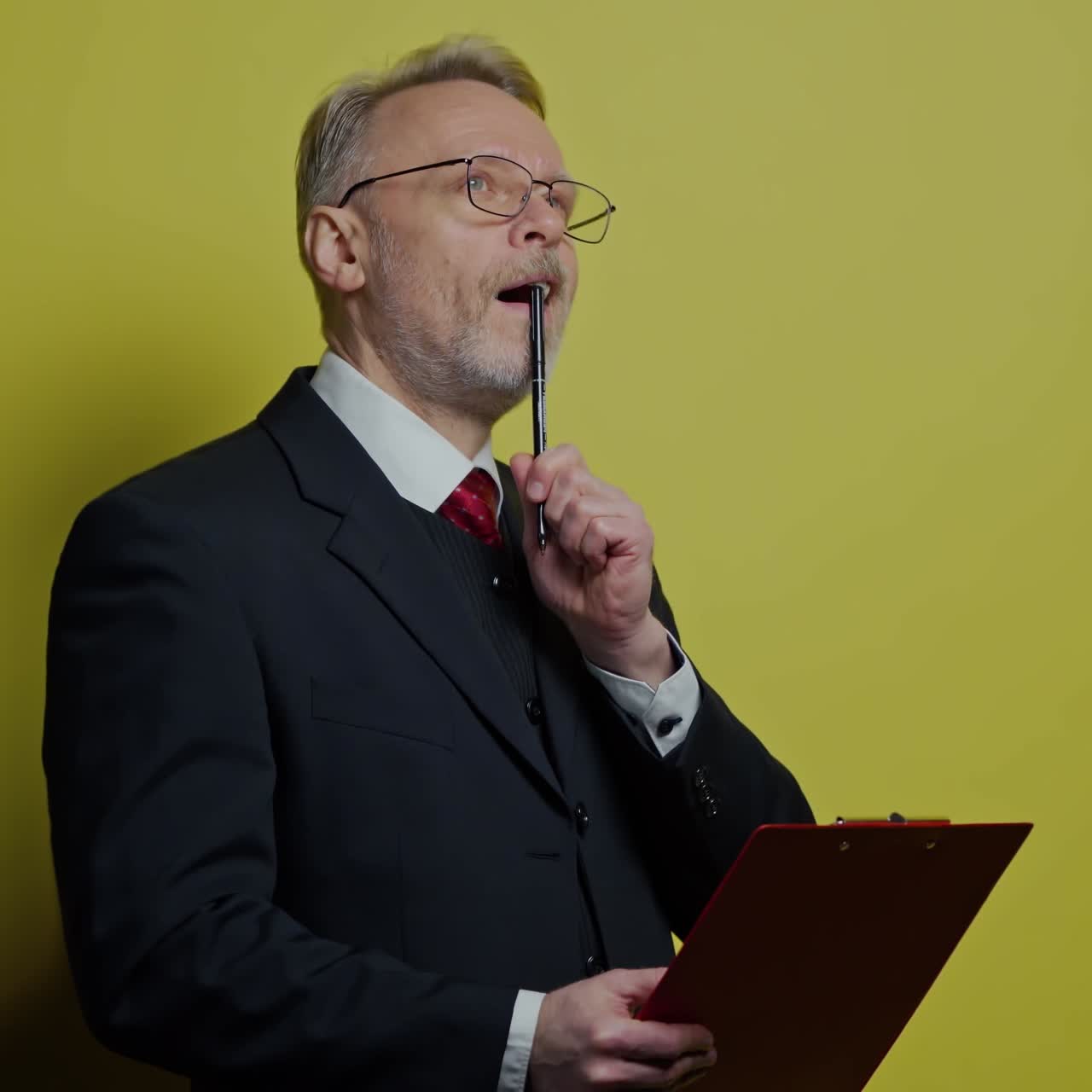 Senior man in costume is writing. Mature businessman in glasses holding a folder and thinking. Portrait of an old professional entrepreneur on yellow background.