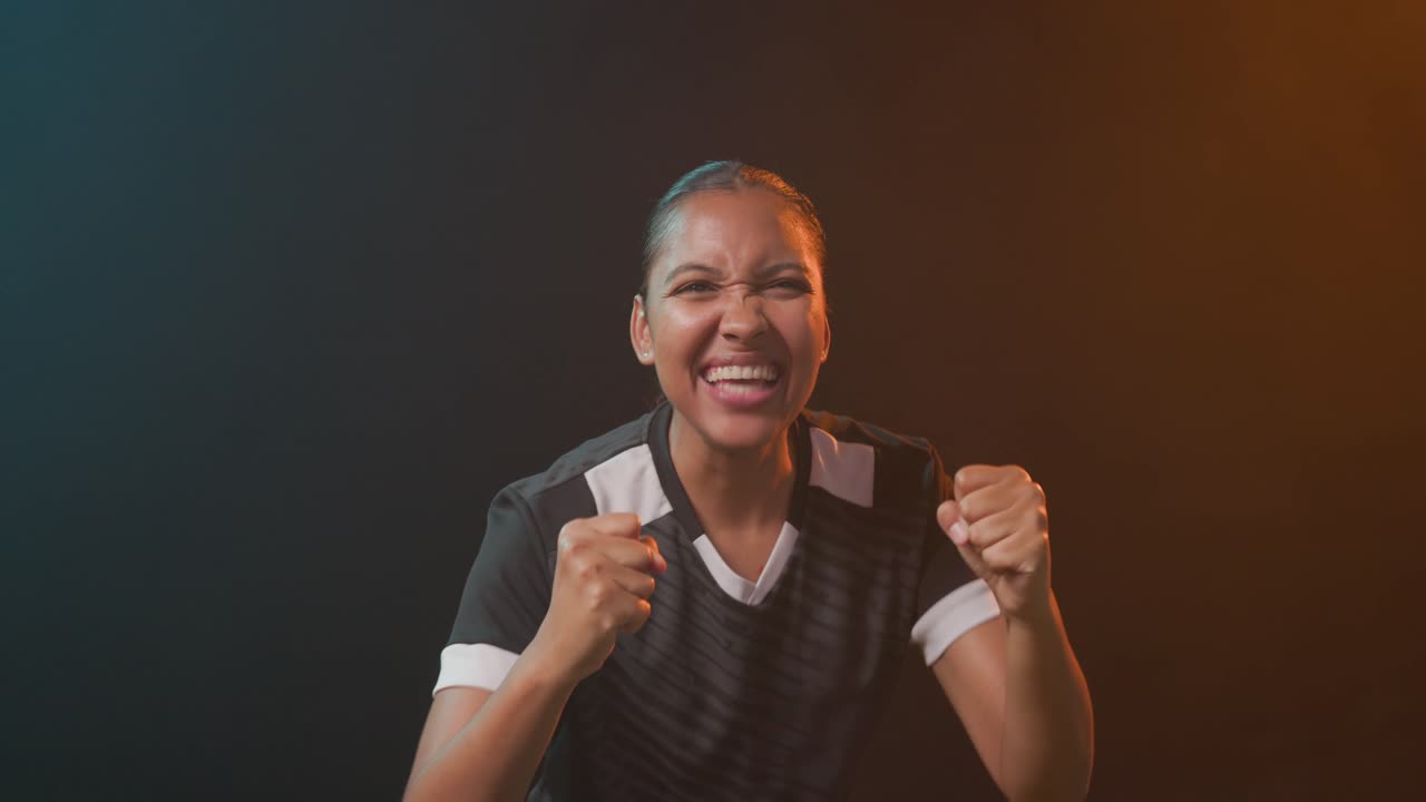 Na revelação, mulher afro-americana celebrando com os punhos erguidos na grama sintética, com uniforme, sob luz azul-petróleo ou laranja.