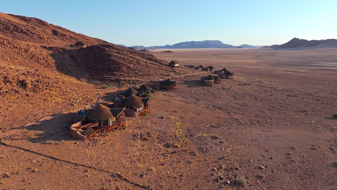 Vista aérea de um acampamento no deserto de chalés de pedra alinhados numa crista nas planícies vermelhas do Namibe ao pôr do sol. O retiro ecológico remoto integra-se na paisagem desértica vermelha sob céus amplos e montanhas distantes.