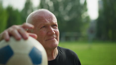 an elderly man in a black top is seen resting his hand on a soccer ball while gazing thoughtfully upward, with blur background of trees and grassy field