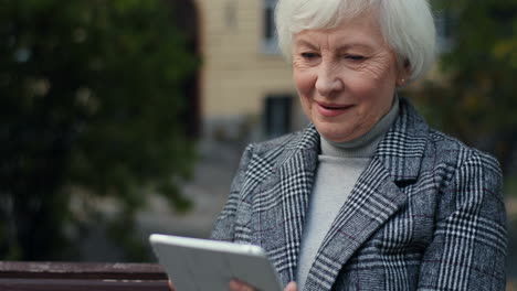 close up view of ederly woman in the coat sitting on the bench in the park and tapping or typing on the tablet in autumn