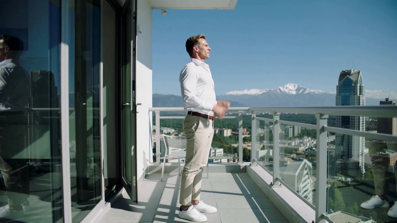 Aprendo la porta a vetri, un uomo in camicia bianca esce sul balcone, sistemandosi la camicia e ammirando le cime.
