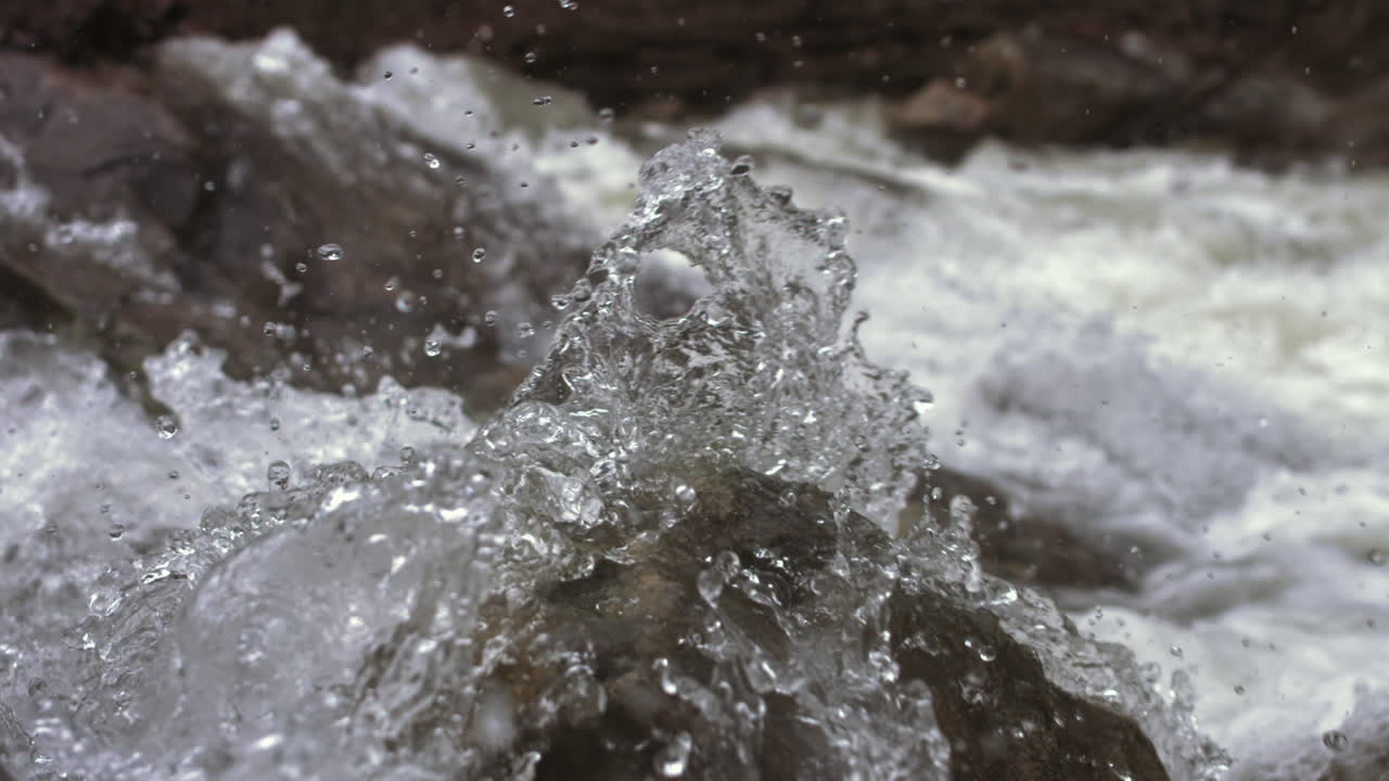 Up close super slow motion footage of droplets of water crashing against the rocks of the rapids of a creek and going up into the air.