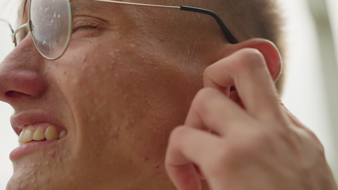 Uomo bianco che si aggiusta gli occhiali vicino all'orecchio, primo piano sulla tempia e sulla pelle umida con goccioline sulla montatura, montature che alternano i ruoli di pendolare, studente, persona che cammina nel parco, lieve disagio e manutenzione ordinaria