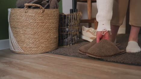 leg view of individual wearing brown crocs walking onto foot mat, removing footwear and placing it into nearby basket, with beige pants, white socks, and wooden interior setting in softly lit room
