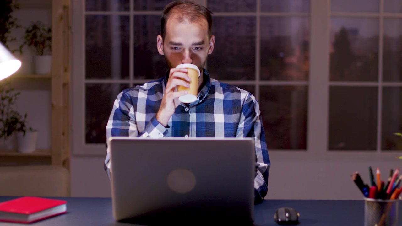 Giovane uomo che lavora al laptop e beve caffè nell'ufficio domestico di notte