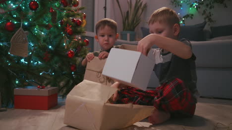 two boys open christmas presents sitting under a decorated christmas tree amid garlands. high quality 4k footage