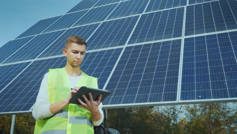 a young engineer in workwear works with a tablet near a large solar panel