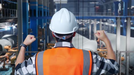 vista cercana de atrás de un ingeniero masculino asiático con casco de seguridad de pie levantando las manos celebrando trabajando en una fábrica de fabricación de turbinas eólicas