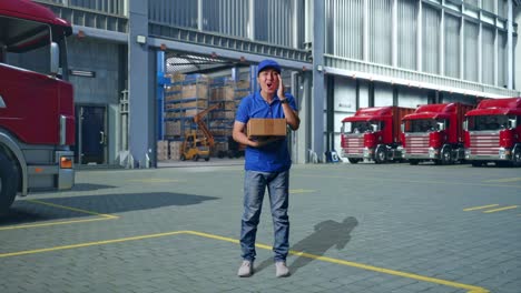 full body of asian male courier in blue uniform smiling and talking to camera while delivering a carton, outside of logistics distributions warehouse
