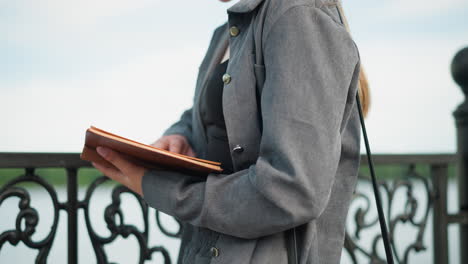 vista parcial de cerca de una mujer sosteniendo un libro abierto con su mano derecha descansando en las páginas y la mano izquierda apoyando debajo de ella, vestida de gris, de pie contra una valla de hierro, leyendo con atención