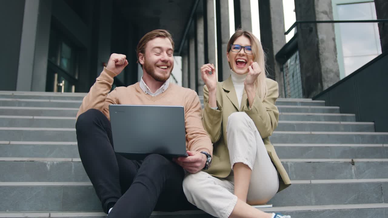 Good-looking positive caucasian young woman and man sitting on steps using laptop, outdoors. Freelancer couple sitting in stairs looking at laptop talking and rejoice in success