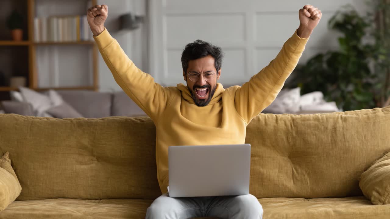 A joyful celebration of triumph unfolds as a man, clad in a cozy yellow hoodie, raises his fists in victory while seated on a plush couch, engaging passionately with his laptop
