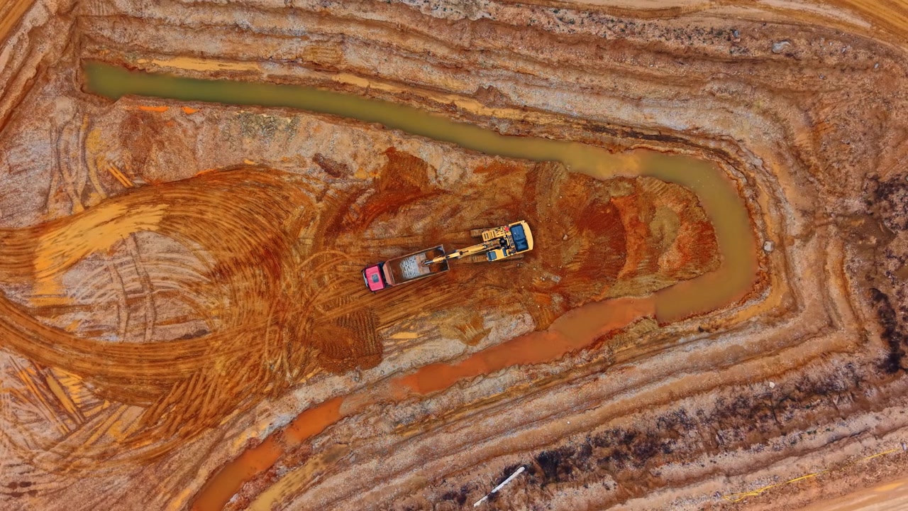 Overhead view of excavator digging in sand quarry. Top down drone shot of yellow machinery amid orange clay, tire tracks and muddy water pools.