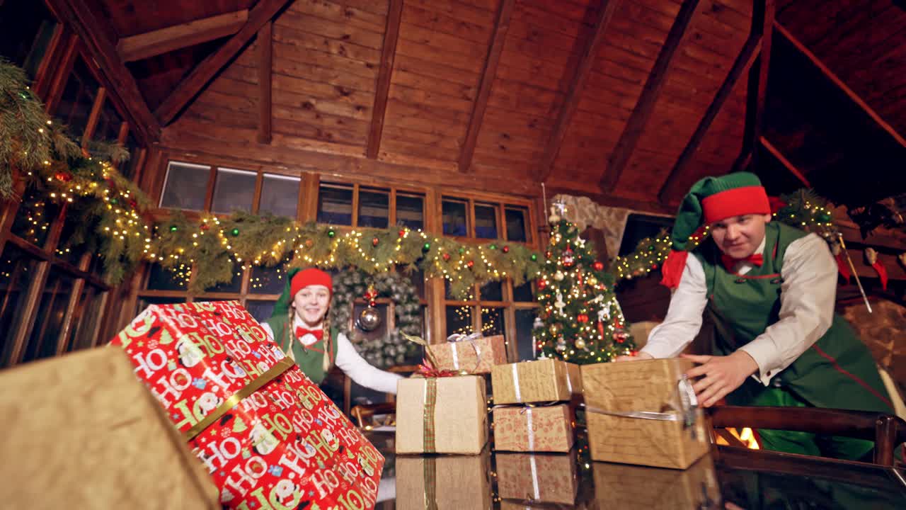 Two elves are standing in specially decorated room near the table, making a piramide from the presents. Feast fireplace in the background. Christmas spirit concept close up video. Christmas eve. New Year is coming. Elves are putting presents on the table. Presents are falling down from the table. El