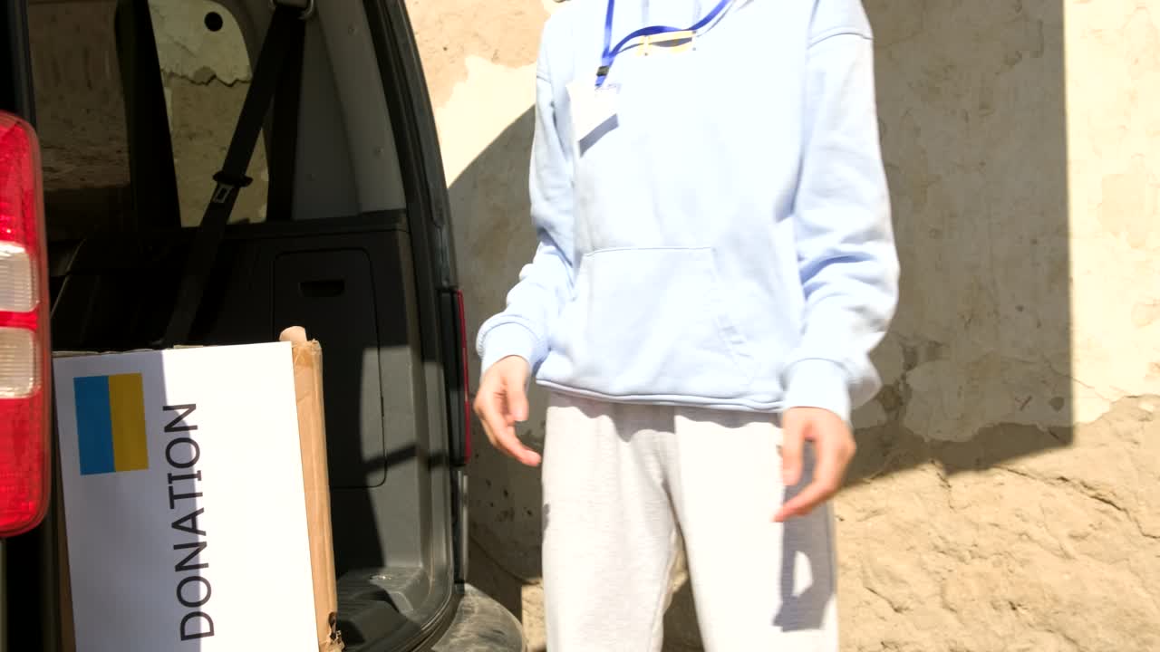 두 명의 자원봉사자 남성과 젊은 여성이 우크라이나 난민들을 위해 기부된 음식과 음료를 담은 자동차 상자에서 내려오고 있습니다.