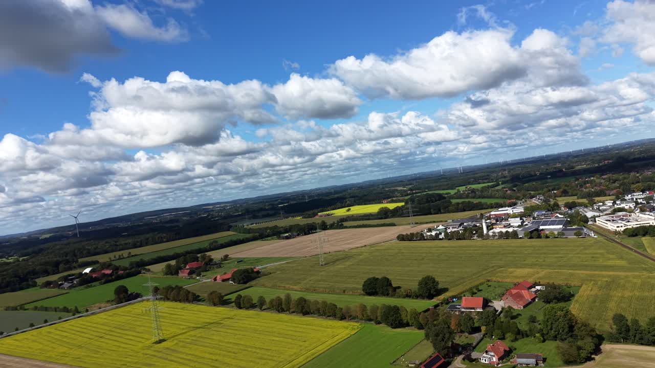 Agricultural colored fields with canola and crops in German countryside. Farmstead with solar panels and industrial district in background. Sunny summer day. Aerial rotating wide shot