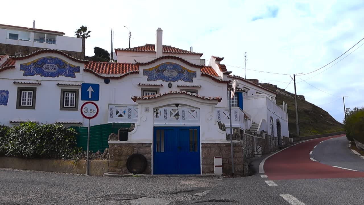 Vista diurna soleggiata di una tradizionale casa portoghese bianca con porte blu, tegole rosse e ceramiche decorative azulejo lungo una tranquilla strada di un villaggio costiero.