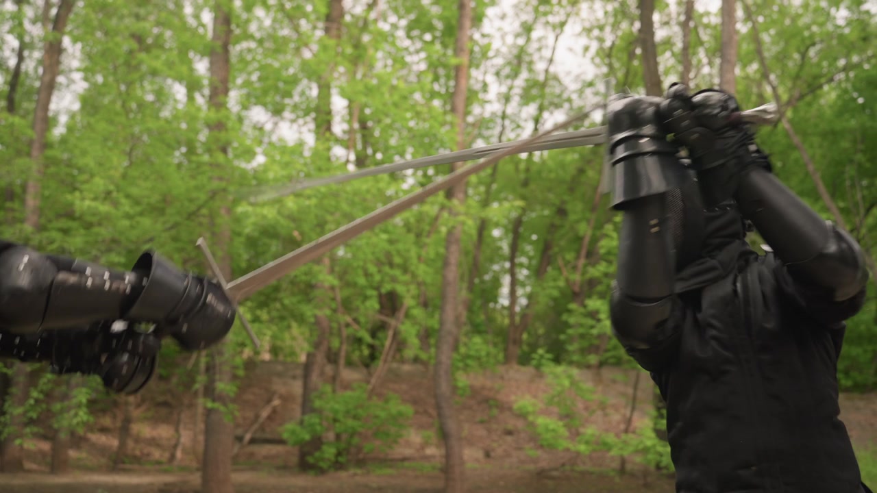 Two people in black fencing gear engage with swords outdoors amidst green trees. One person holds sword while other parries both wearing protective masks and gloves. scene features close