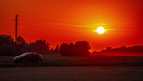 Un-Coche-Aparcado-A-Lo-Largo-De-Una-Carretera-Rural-Mientras-Un-Sol-Ardiente-Desciende-Detrás-De-Una-Capa-De-árboles---Lapso-De-Tiempo