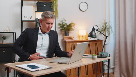 sick young businessman in suit using laptop suddenly clutching stomach in pain at home office desk