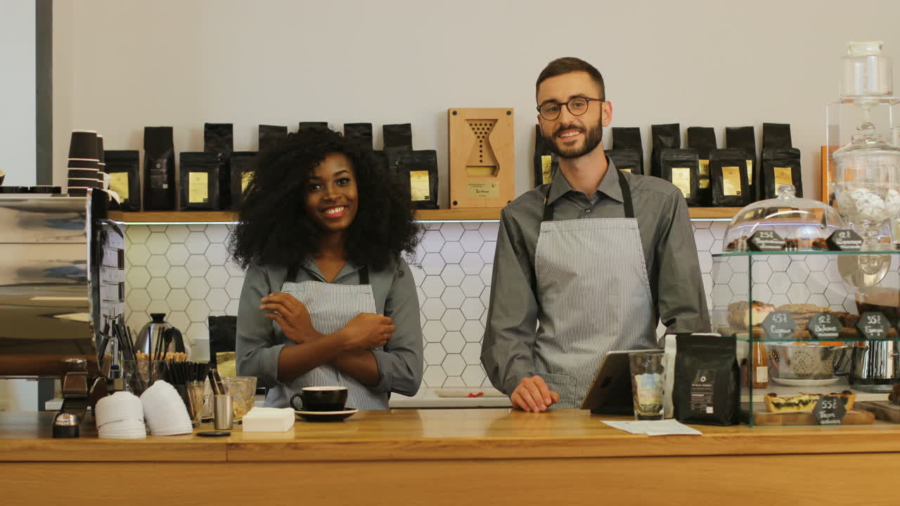 barista afroamericana y su compañero de trabajo sonriendo y mirando la cámara con los brazos cruzados detrás de la barra de la cafetería