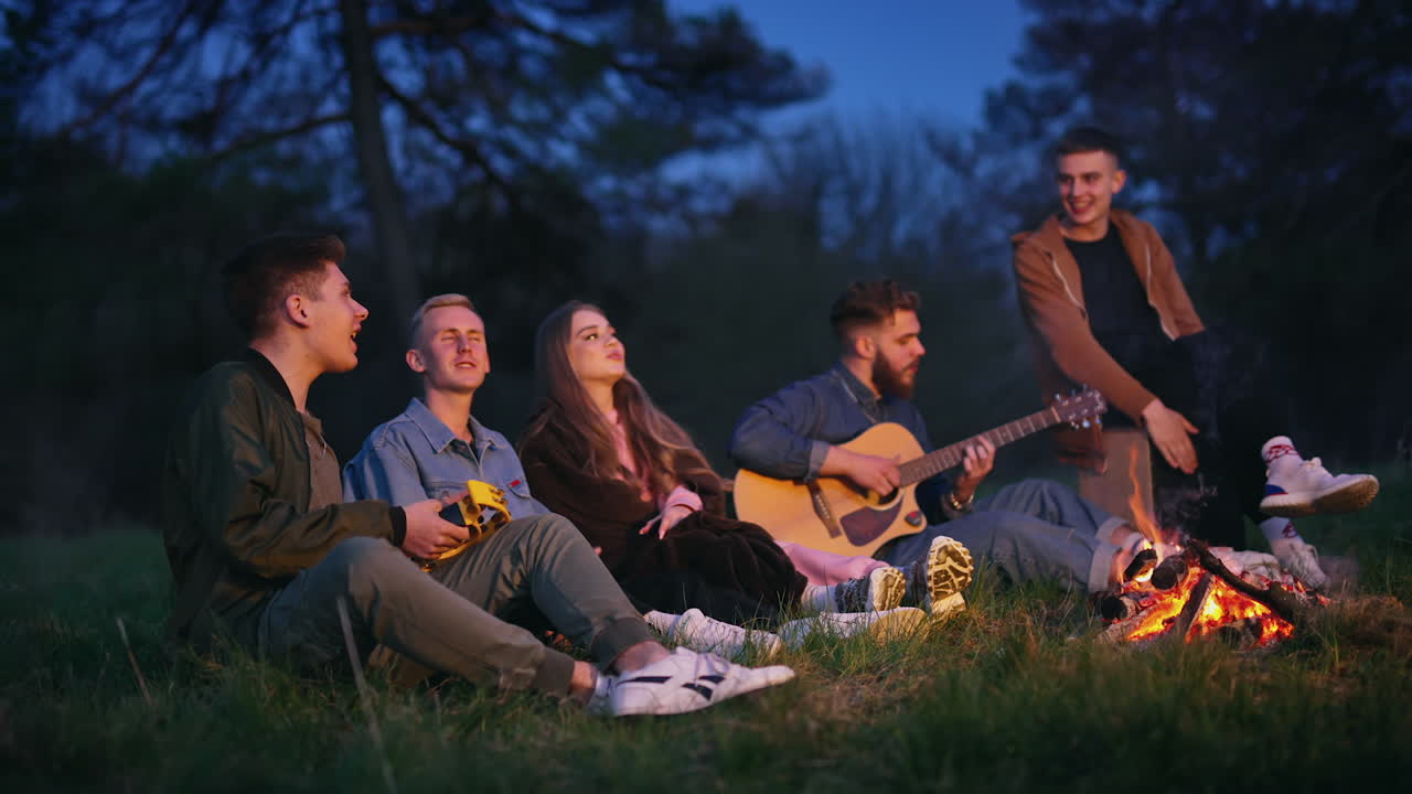 Happy young friends singing together. Young men and a beautiful girl sitting near the fire among nature and spend time to the guitar music at night.