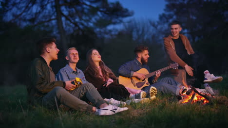happy young friends singing together. young men and a beautiful girl sitting near the fire among nature and spend time to the guitar music at night.