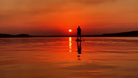 orange sun setting sky over the calm river. man floats on a sup board using a paddle.