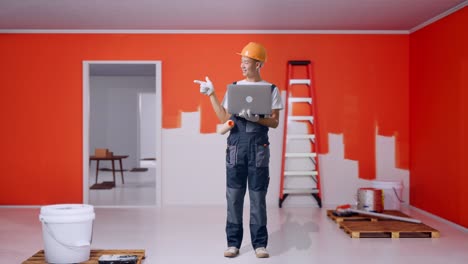 full body of asian man painter wearing safety helmet using a laptop and pointing to side while standing with painting wall in room