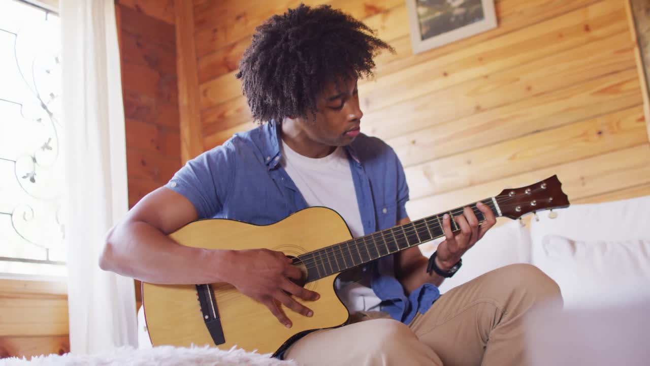 Glad afroamerikansk mand sidder på sofaen og spiller guitar i en bjælkehytte, slow motion