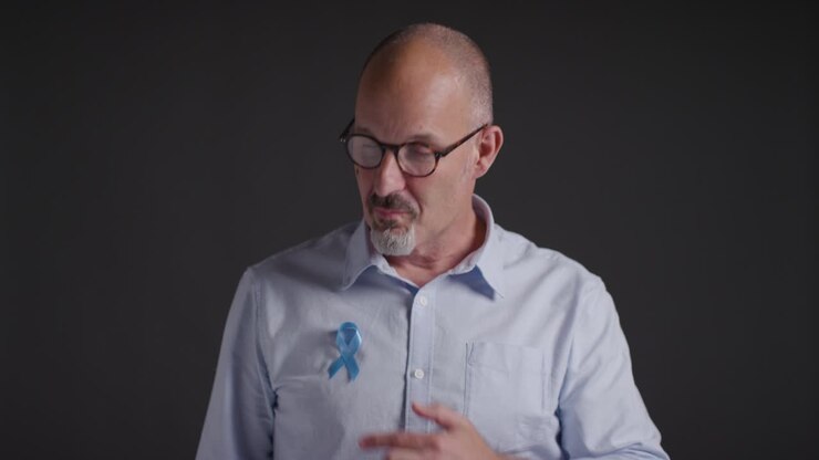 Portrait Of Mature Man Putting On Blue Ribbon Badge Symbolizing Awareness Of Men&#039;s Health And Cancer Onto Shirt