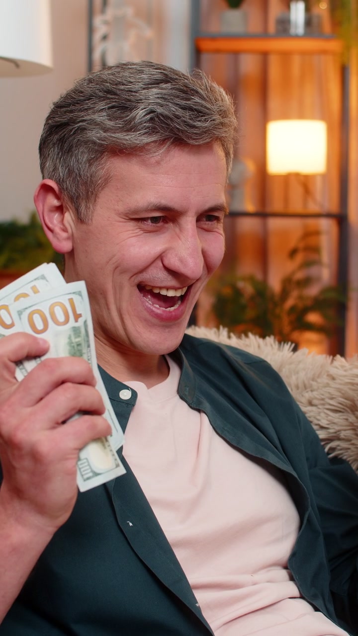 Jonge man lacht terwijl hij contante dollars in zijn handen telt, wat vreugde uitdrukt over inkomen en financieel gewin.