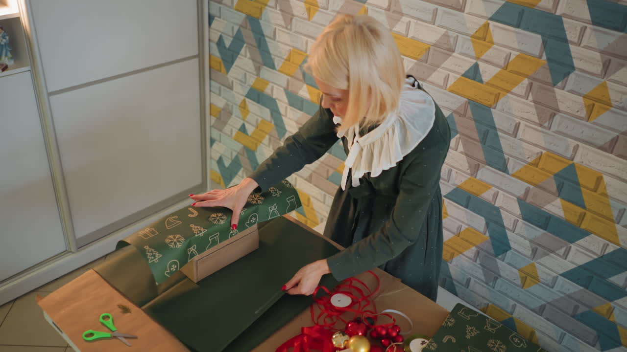 Donna bianca che incarta con perizia un grande regalo di Natale su un bancone, srotolando carta verde, levigando i bordi, fissando nastro adesivo e fiocco, parete luminosa a fantasia, flusso di lavoro efficiente e concentrato, organizzazione da negozio.