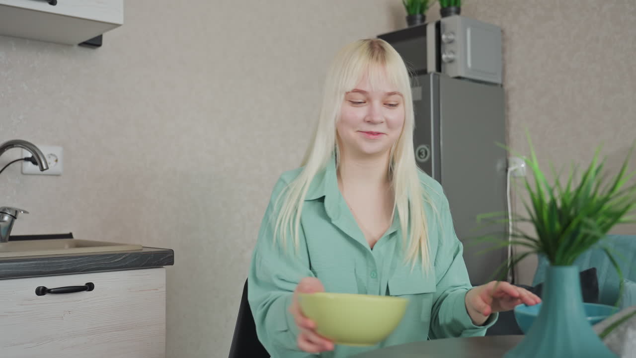 young woman seated in kitchen happily drops bowl of different size on table, modern home with kitchen appliances, decorative plant, fridge, and cozy interior visible