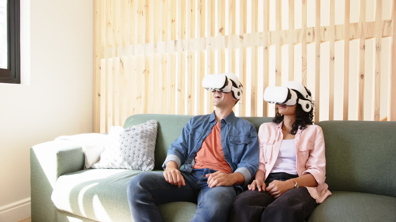 Using VR headsets, young multiracial couple sitting on couch and pointing at screen, at home