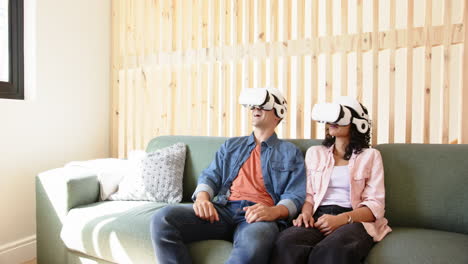 using vr headsets, young multiracial couple sitting on couch and pointing at screen, at home