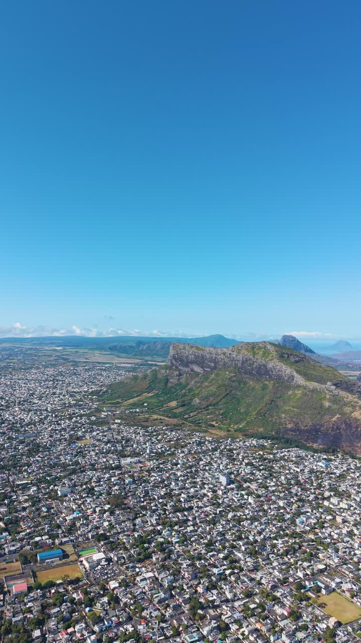 Vertical aerial view of Beau Bassin–Rose Hill, Mauritius, featuring dramatic mountain ridges rising above the urban landscape under a clear tropical sky