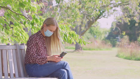 Una-Pareja-Socialmente-Distanciada-Con-Máscaras-Se-Reúne-En-El-Parque-Durante-Una-Pandemia-De-Salud-Mirando-Teléfonos-Móviles