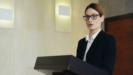 close-up view of caucasian businesswoman speaker on a podium wearing eyeglasses and formal clothes and talking in a conference room