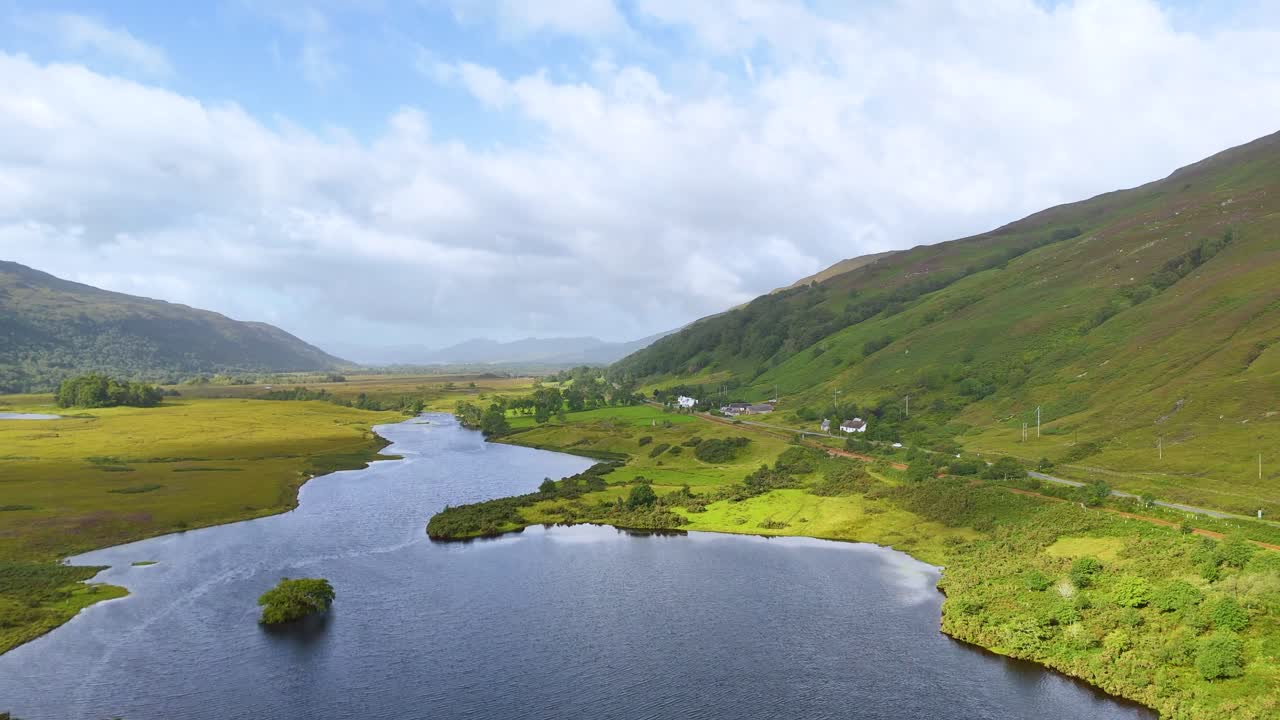 Aerial camera smoothly glides above a winding river, grassy fields, and green hills under partly cloudy daylight in the Scottish Highlands