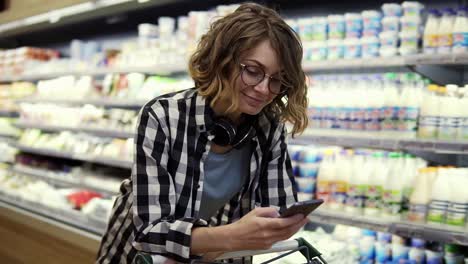 compras, tecnología, ventas, consumismo y concepto de personas - mujer con teléfono inteligente y auriculares en el cuello caminando en el supermercado