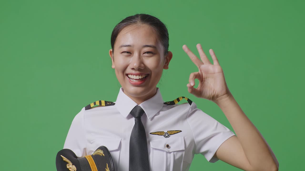 グリーンバックのスタジオで、笑顔でカメラにOKサインを見せるアジア人女性パイロットのクローズアップ